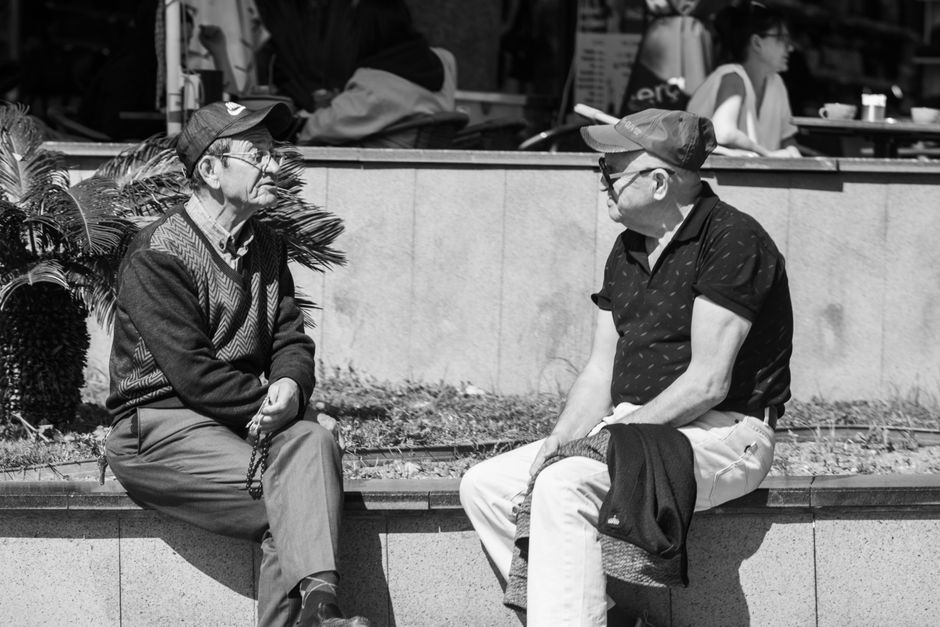 晴れた日に、都会の屋外で二人の高齢男性が座って談笑している。