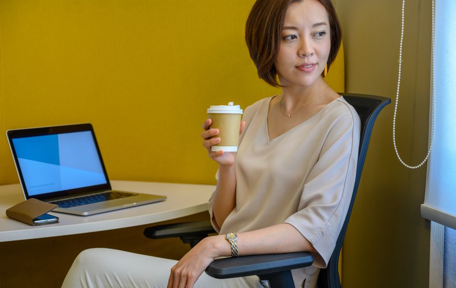 ラップトップとコーヒーを使っているデスクに座っている女性が、仕事中のリラックスした瞬間を楽しんでいます。