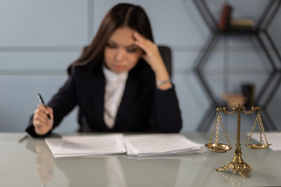 近くに正義の天秤がある疲れた女性弁護士が書類を確認している様子が、法律業務におけるストレスを強調しています。